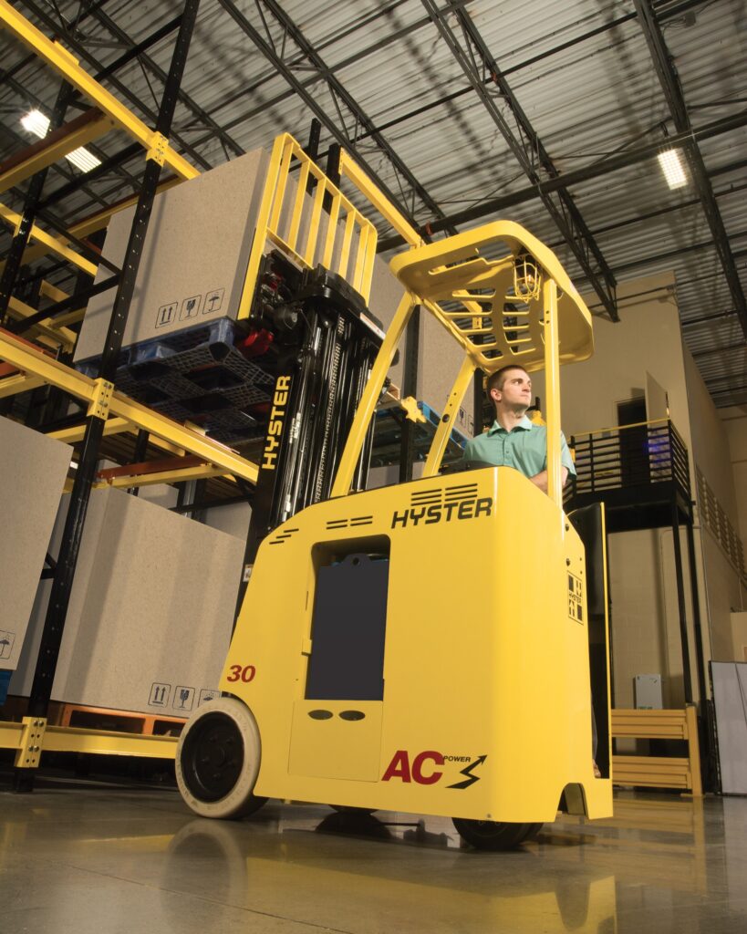 Photo of a man operating a Hyster stand-up 3-wheel electric forklift in a warehouse.